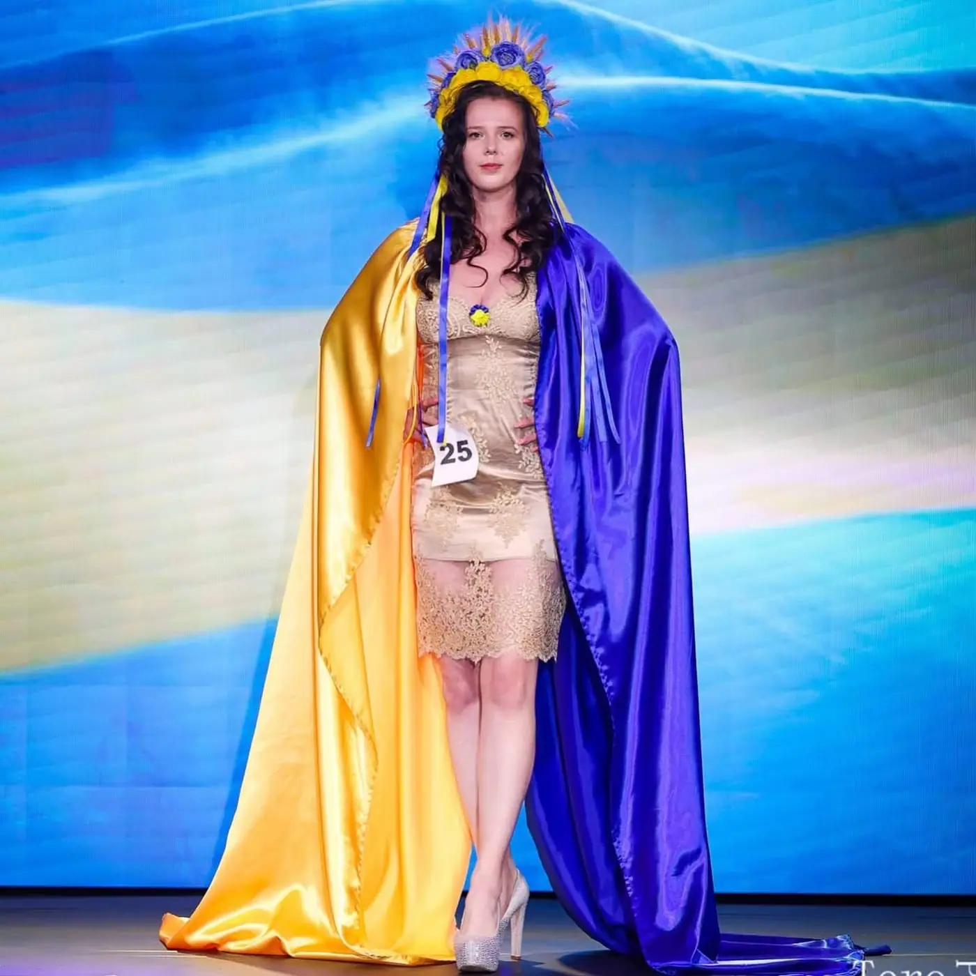 A contestant on stage in a blue and yellow flag cape and golden headdress