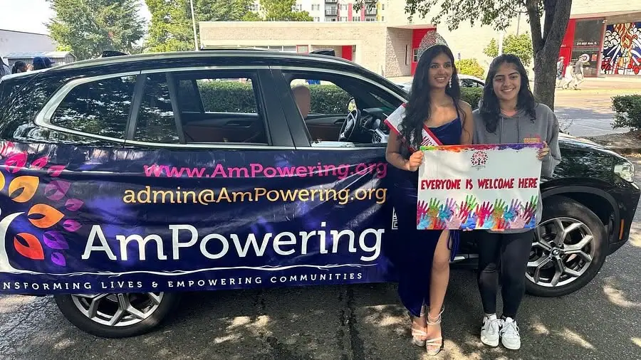 A titleholder and a volunteer beside the AmPowering car, holding an “Everyone is welcome here” sign