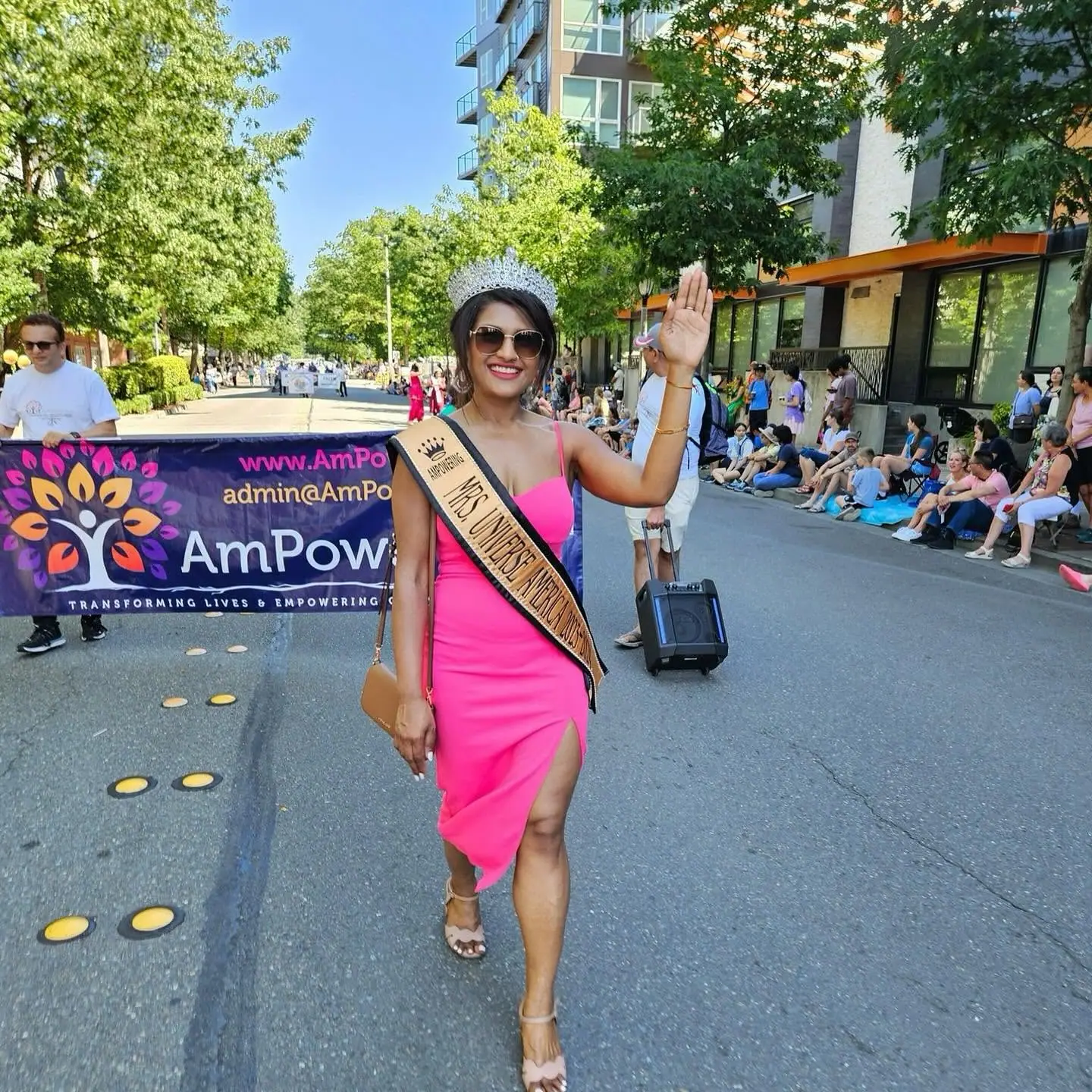 Mrs Universe America in a pink dress waving as she walks ahead of the AmPowering banner in a parade