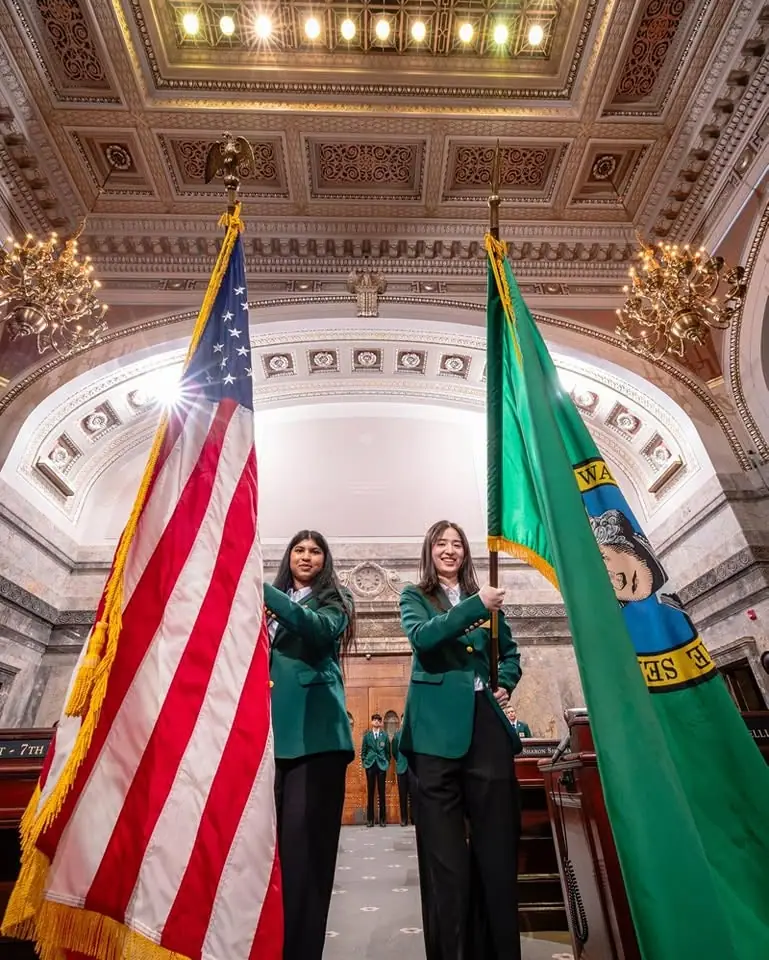 Two Senate pages in green blazers carrying the US and Washington State flags in the Senate chamber