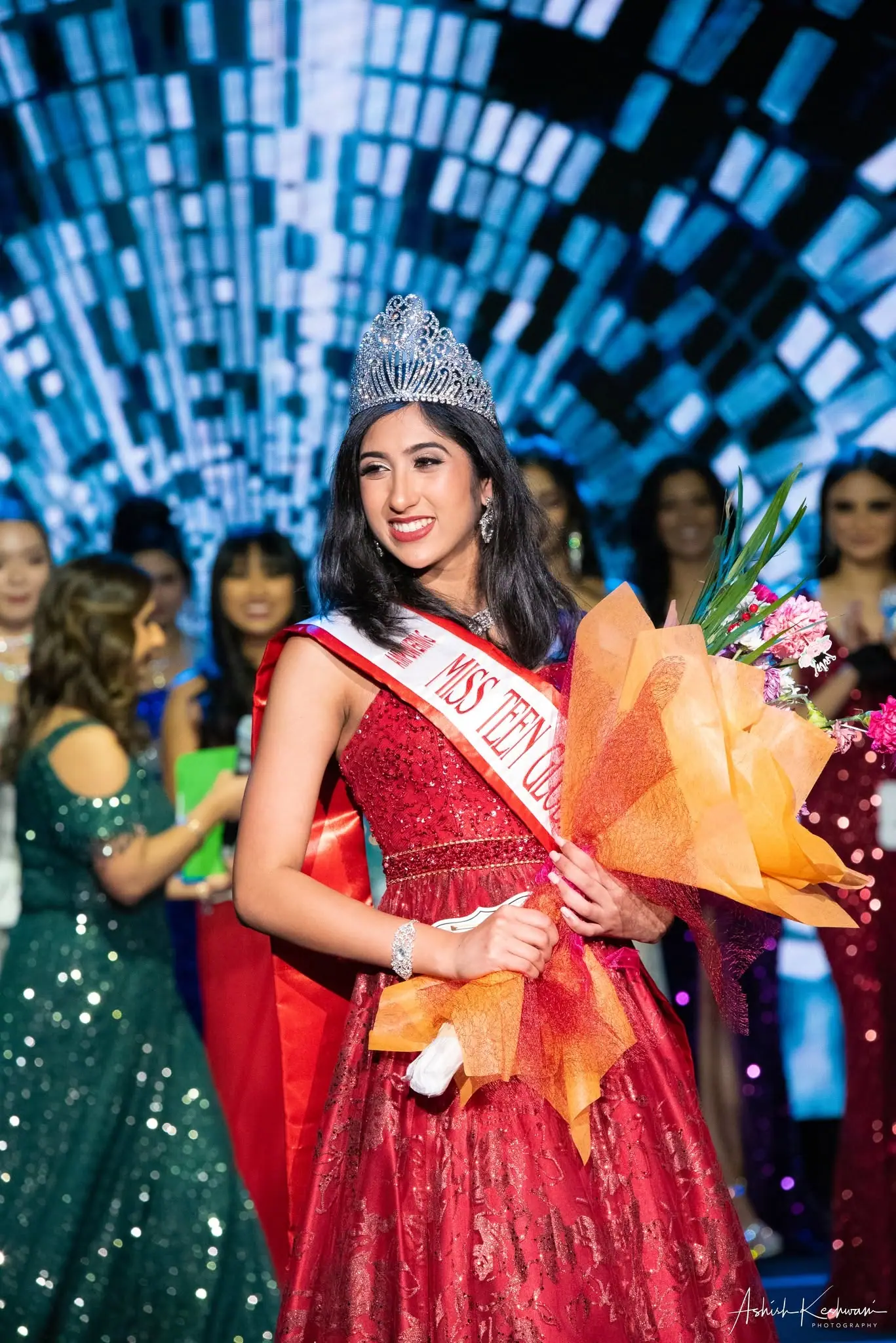 The newly crowned Miss Teen Global Asia in a red gown, holding flowers on stage