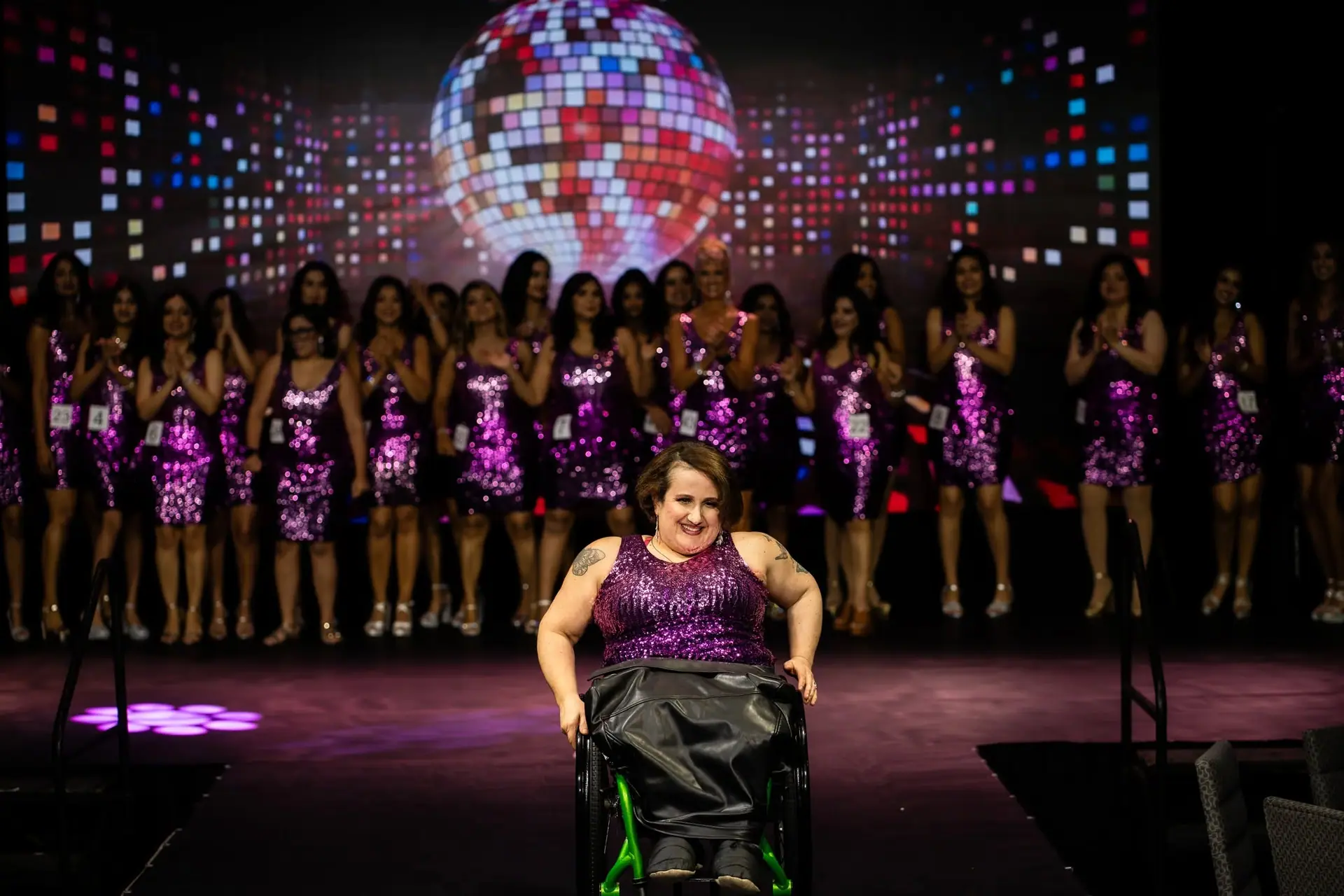 A contestant in a wheelchair smiling on the runway as the other contestants applaud behind her